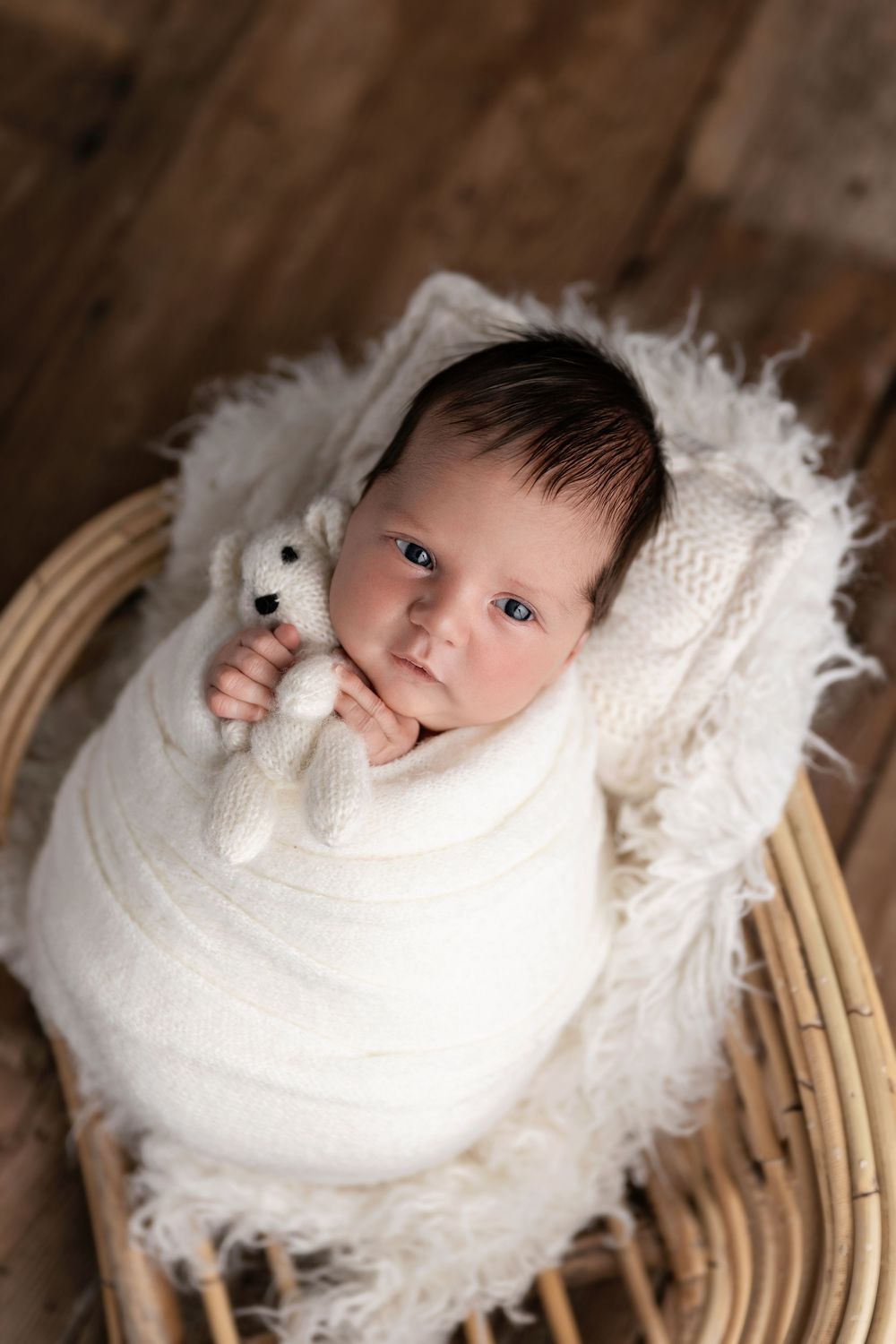 Newborn baby swaddled in white wrap holding a knit teddy bear, resting in a wicker basket with fluffy white lining.