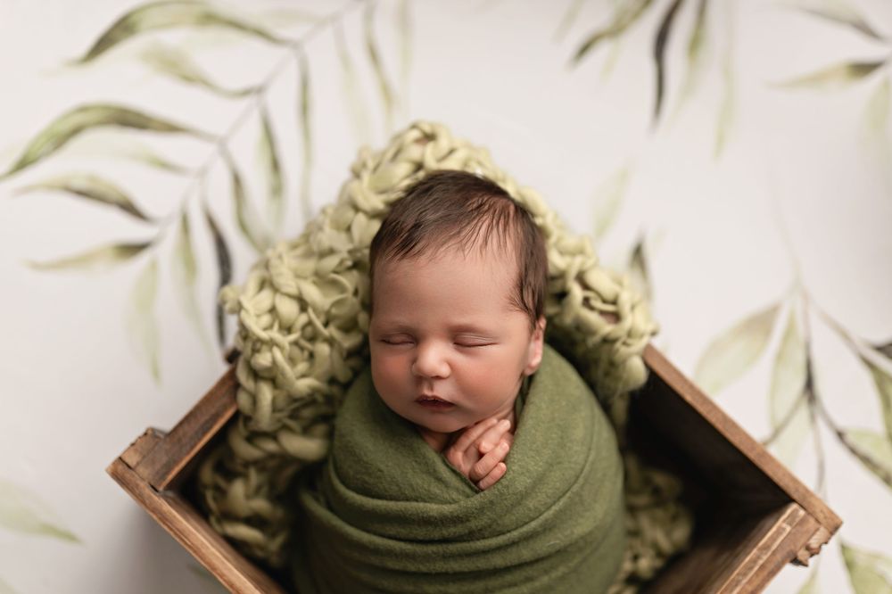 Sleeping newborn baby wrapped in green swaddle nestled in wooden box with leaf backdrop.
