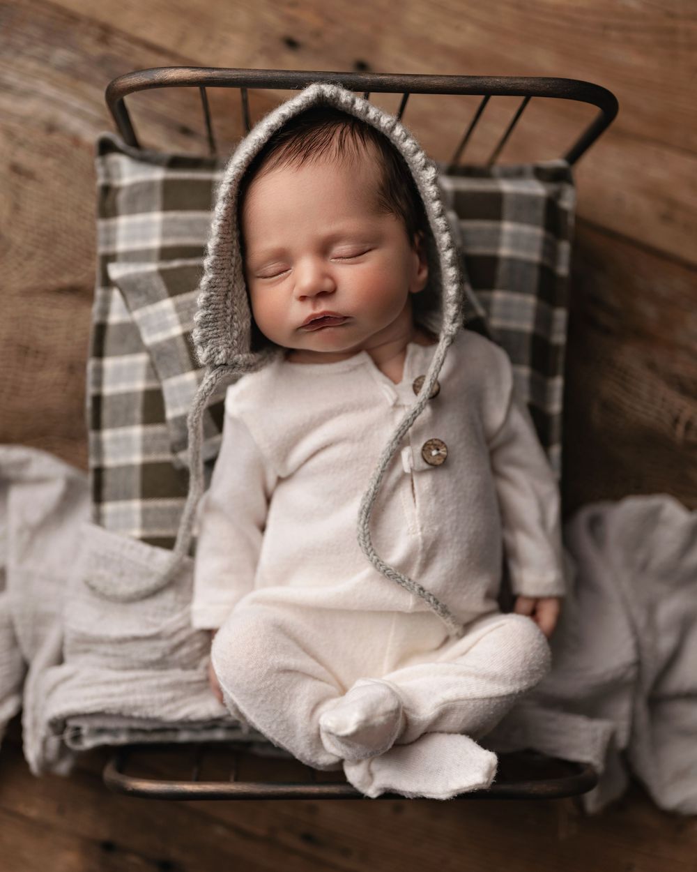 Sleeping newborn baby in white romper and gray bonnet resting in a small vintage metal bed with plaid blanket.