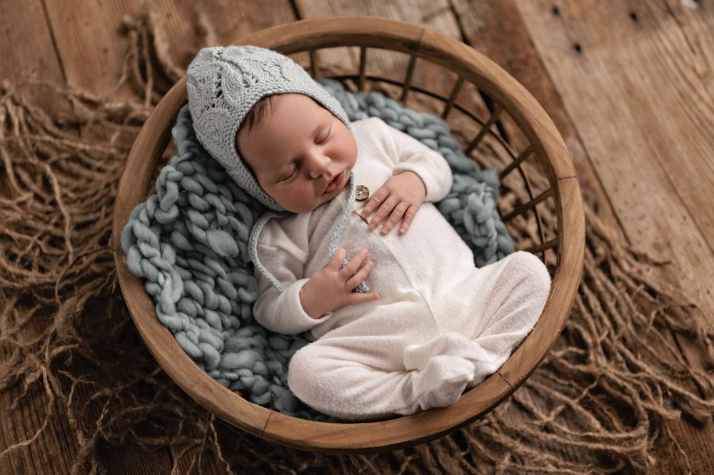 Newborn baby sleeping peacefully in a wooden basket with blue knit blanket and matching hat.