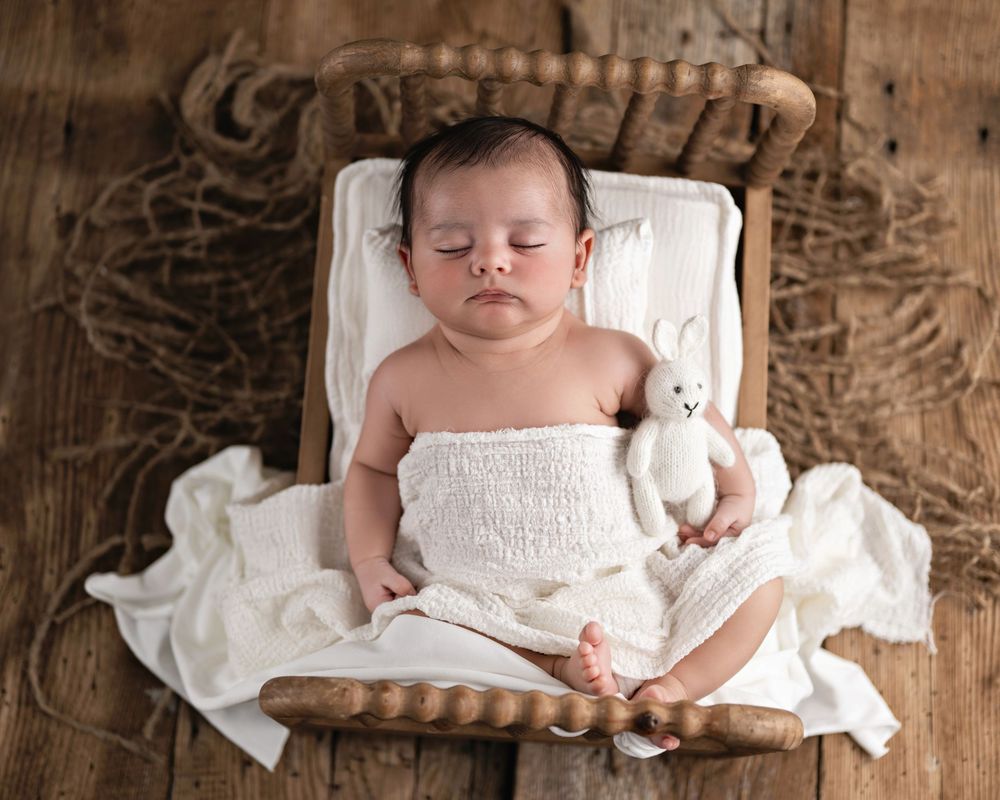 Sleeping newborn baby wrapped in white linen on a rustic wooden mini bed holding a small white bunny toy.