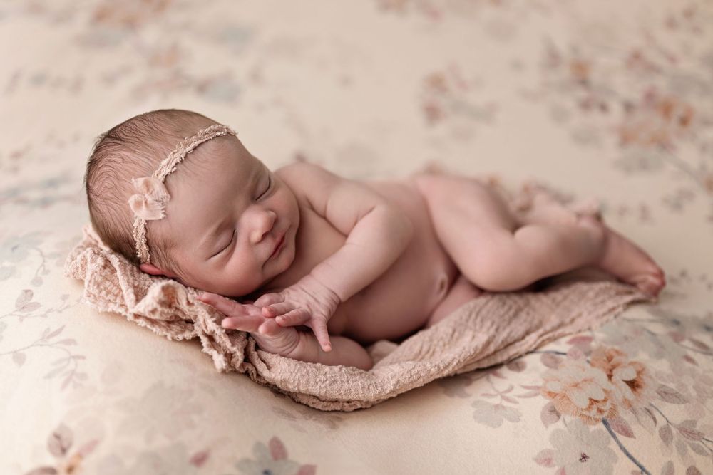 Sleeping newborn baby wearing delicate headband rests peacefully on soft beige blanket with floral pattern.