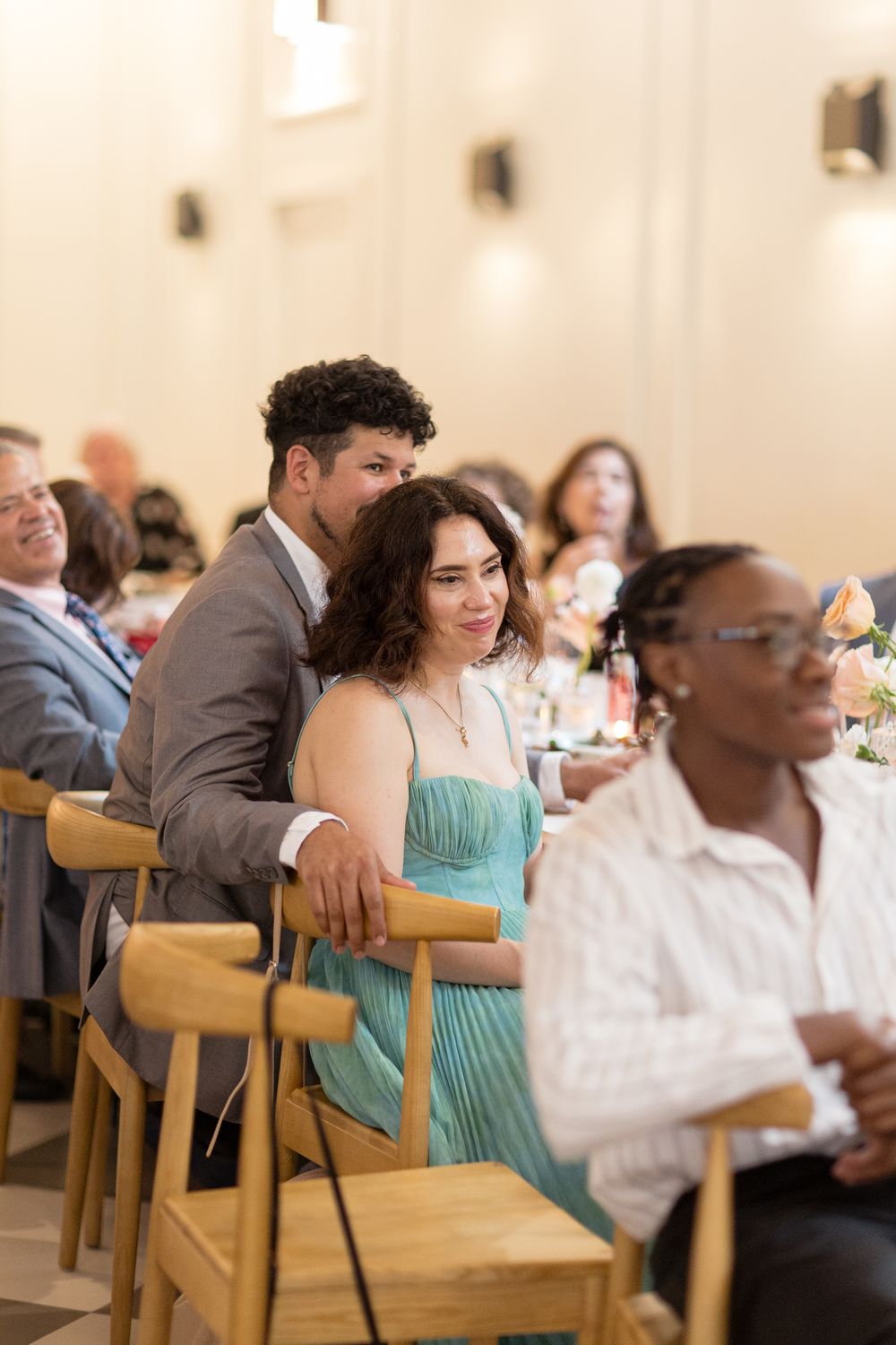 Wedding guests enjoy a reception dinner while seated at wooden chairs in an elegant light-filled venue.