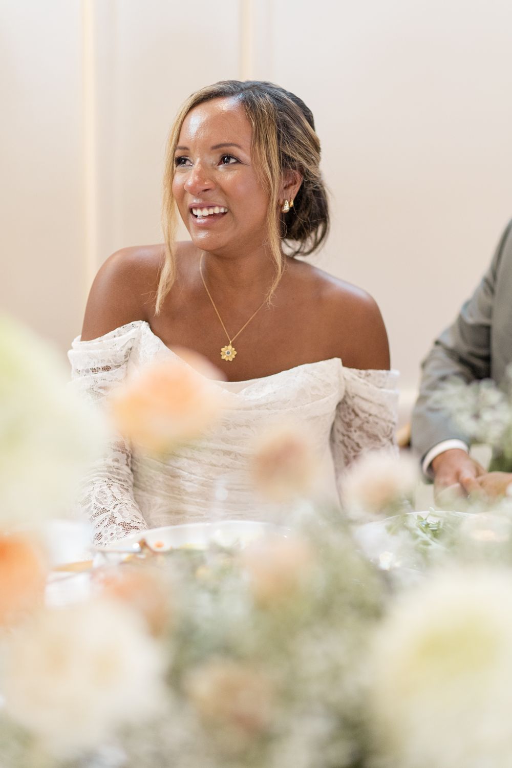 Bride smiling joyfully while seated at wedding reception table looking up with thoughtful expression.