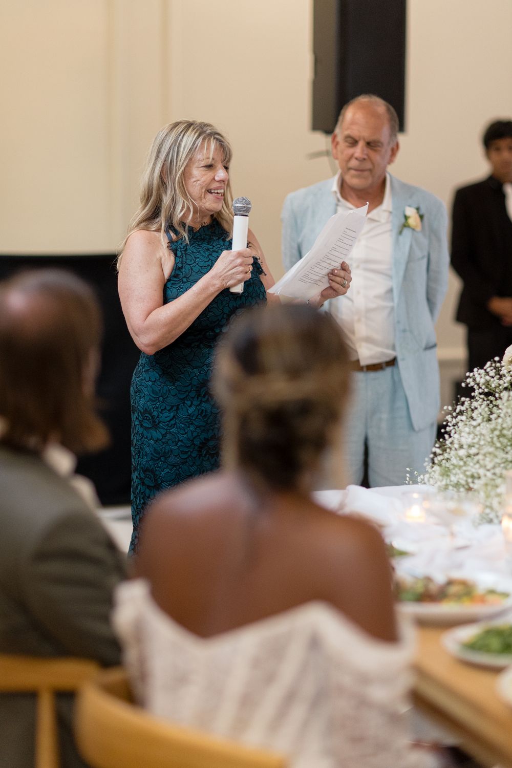 Wedding guests share toasts and speeches during reception dinner at tables decorated with white flowers.