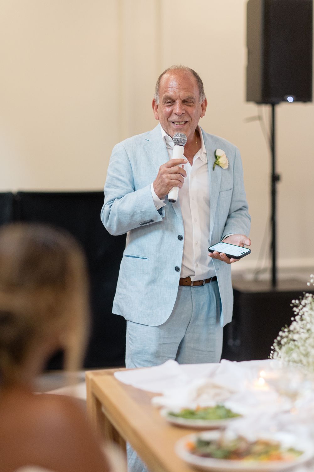 A person in a light blue jacket giving a speech at a wedding reception while holding a drink.