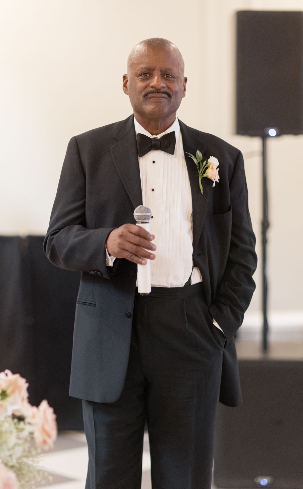 A wedding guest in a black tuxedo and bowtie speaks into a microphone while wearing a white boutonniere.