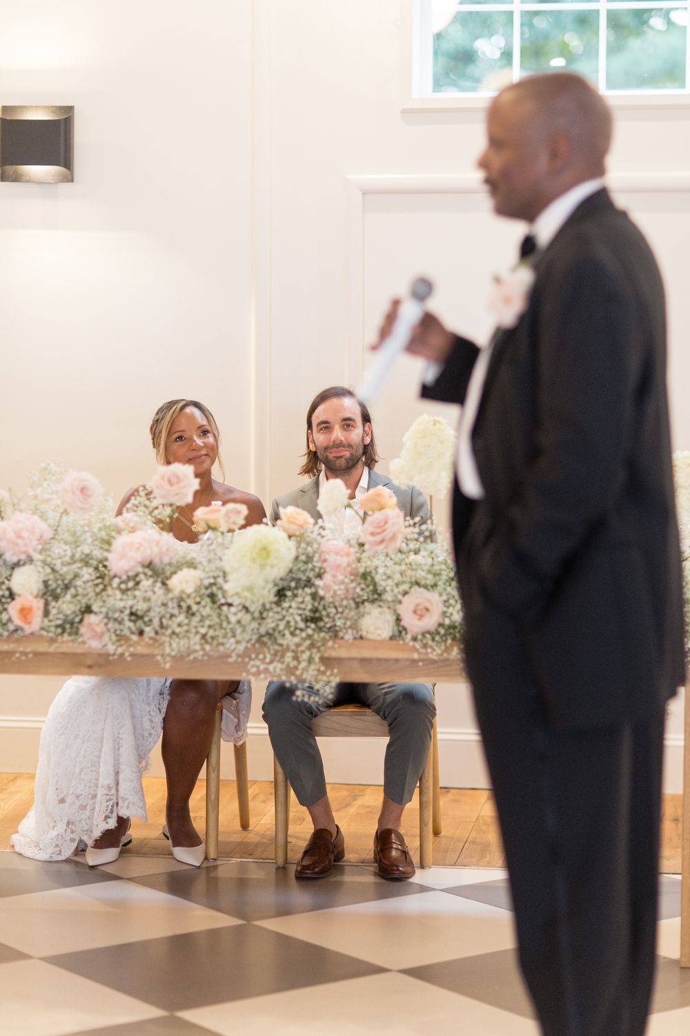 Guests seated at a wedding reception with elegant floral arrangements of baby's breath and pink roses on long wooden tables.