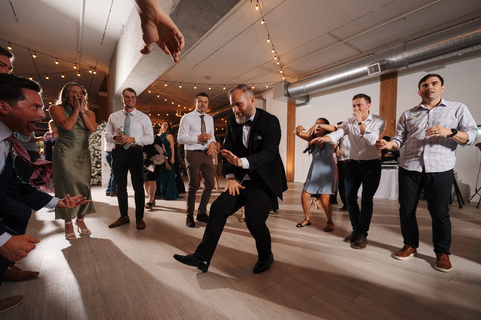 Wedding guests dance together on the dance floor at an indoor reception with string lights overhead.