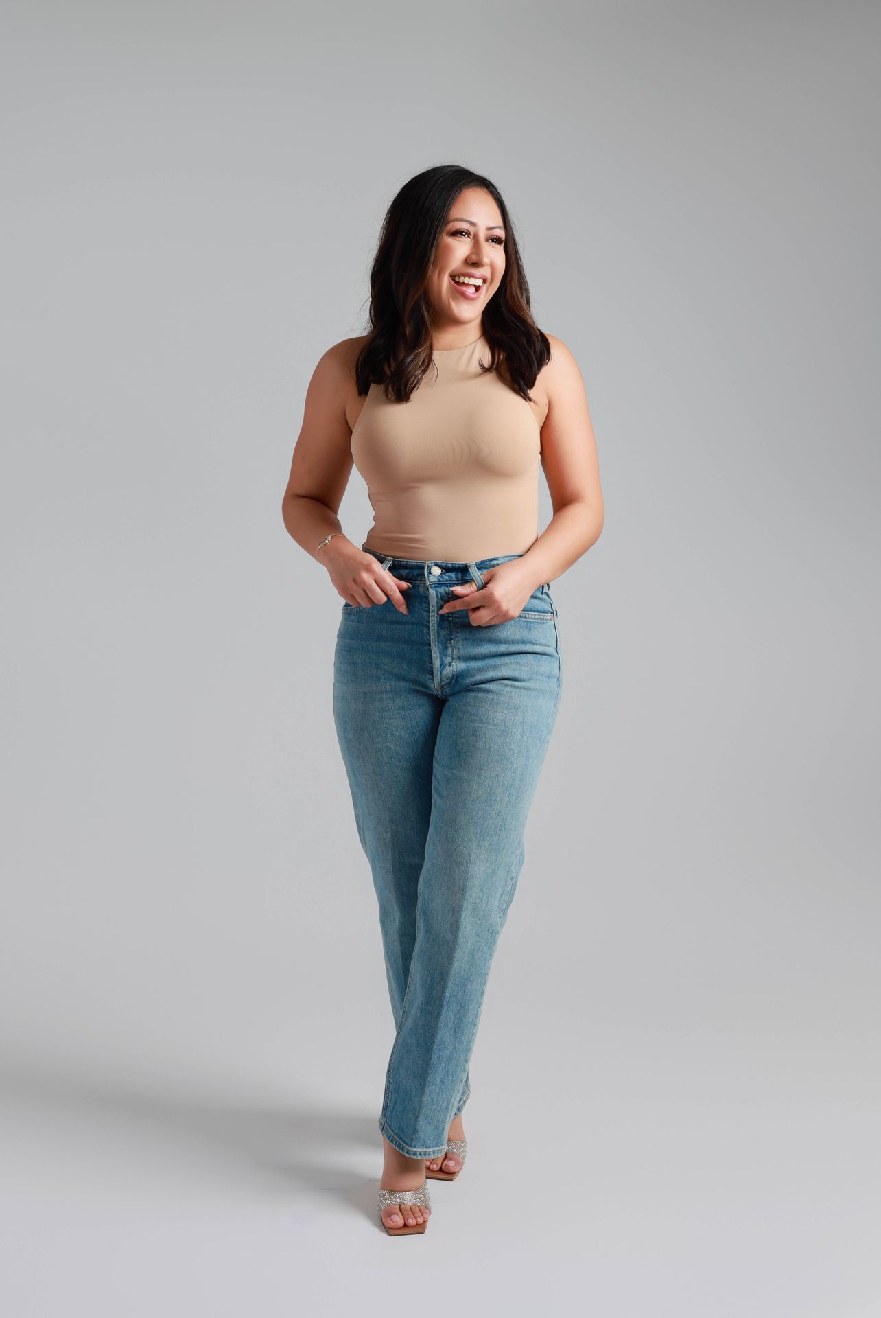 A series of photos showing someone in a beige top and light wash denim jeans posing against a grey backdrop.
