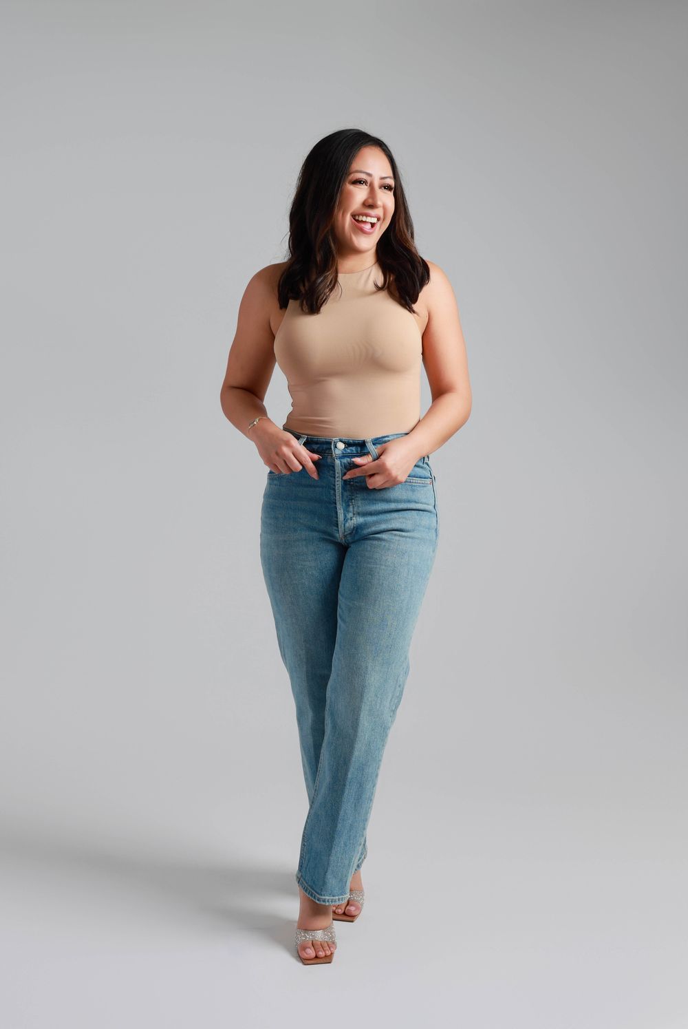 A series of photos showing someone in a beige top and light wash denim jeans posing against a grey backdrop.