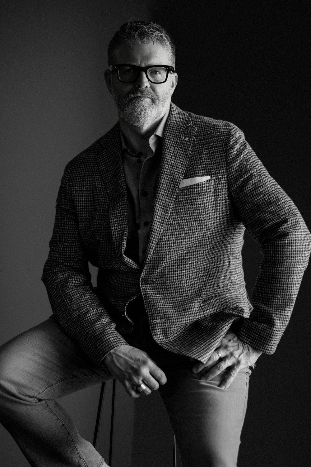 Black and white portrait of a distinguished middle-aged man with gray beard, glasses, and blazer seated confidently.