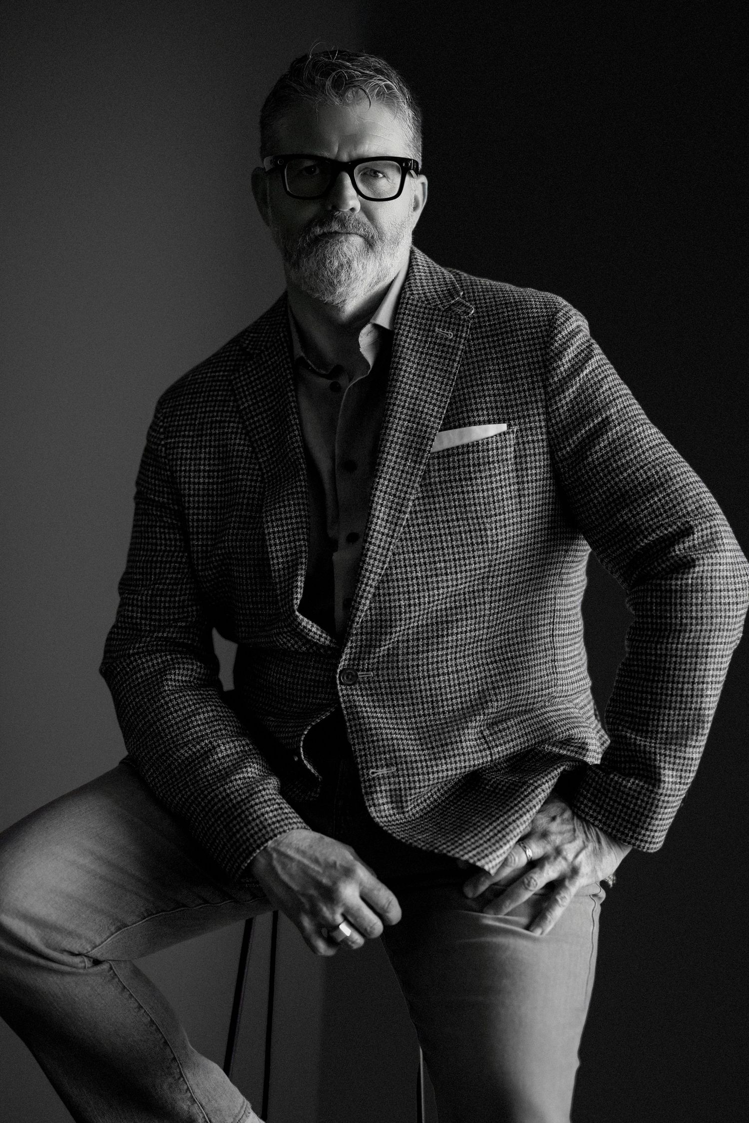 Black and white portrait of a distinguished middle-aged man with gray beard, glasses, and blazer seated confidently.
