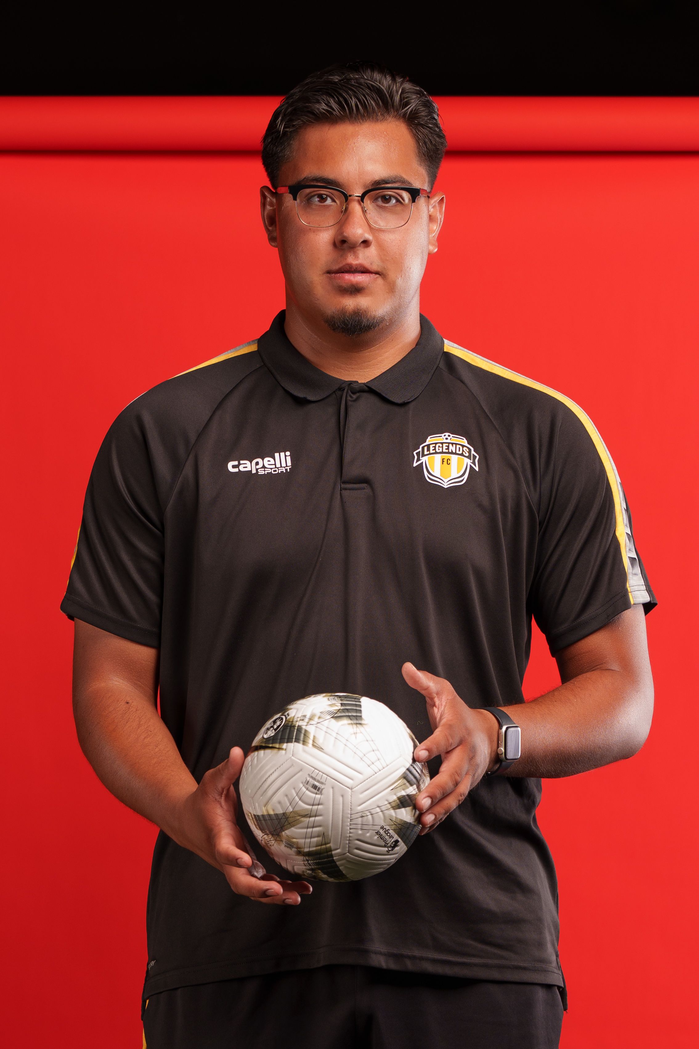 Soccer coach in black polo uniform holds white ball against red background.