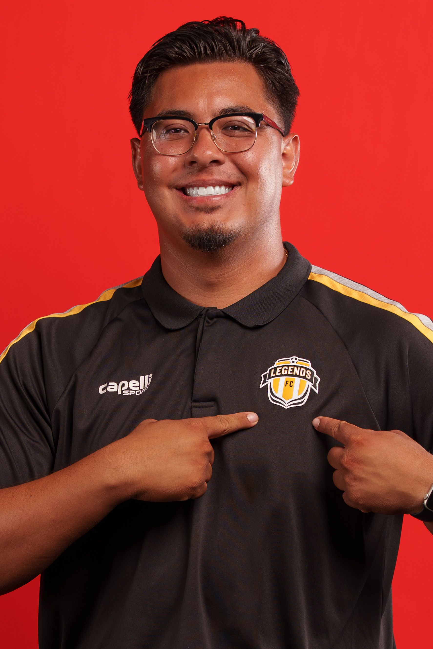 Coach in black polo points to badge logo on uniform against red background.
