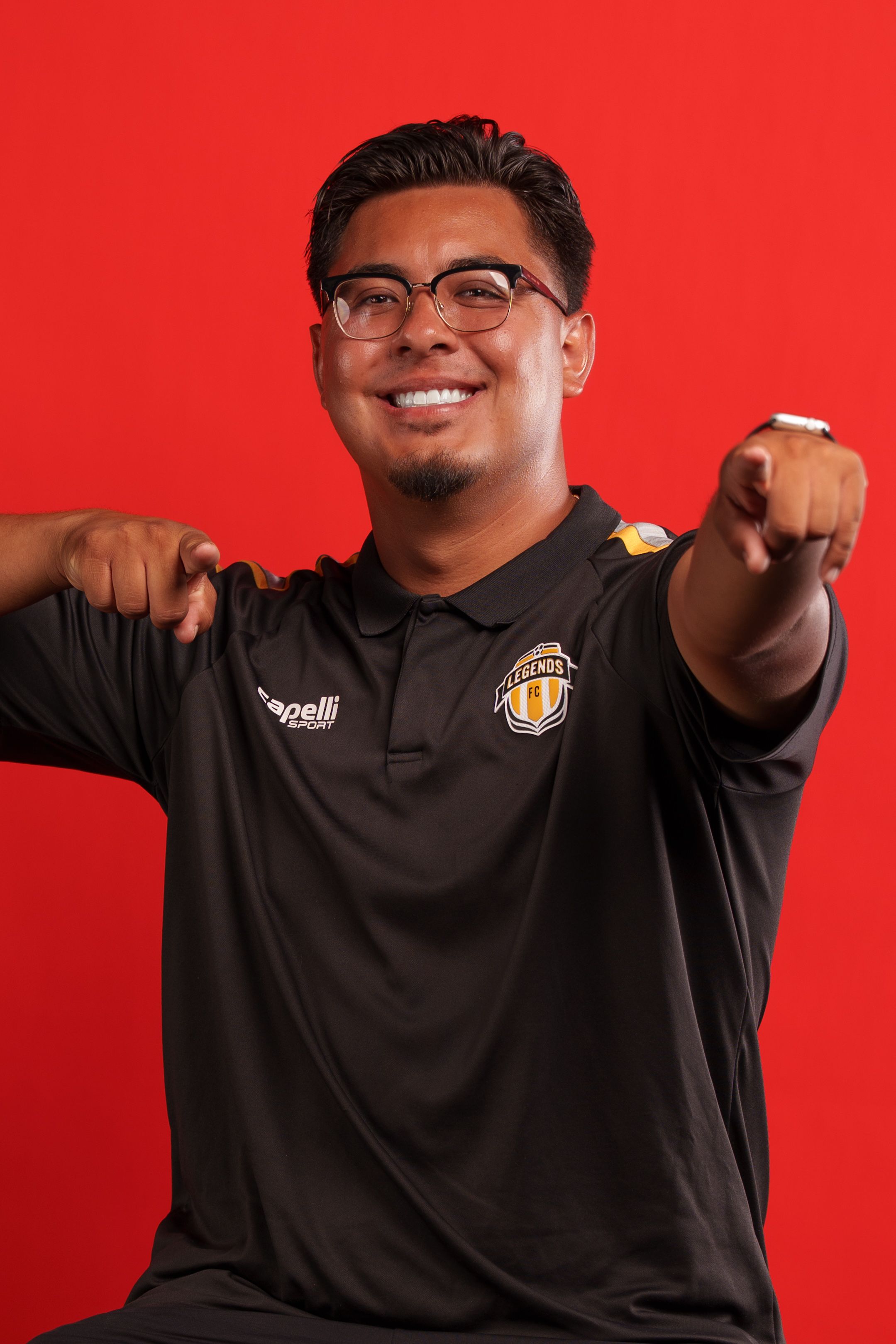 Coach gestures enthusiastically in black uniform against red studio backdrop.