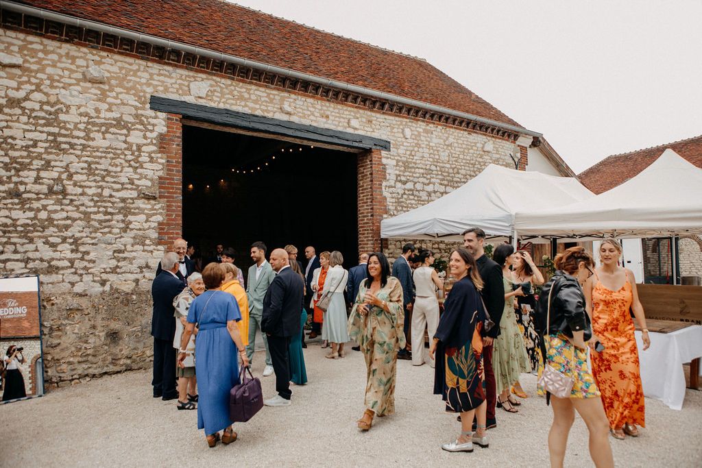 Wedding guests mingling outside rustic stone barn venue with string lights and white tent.