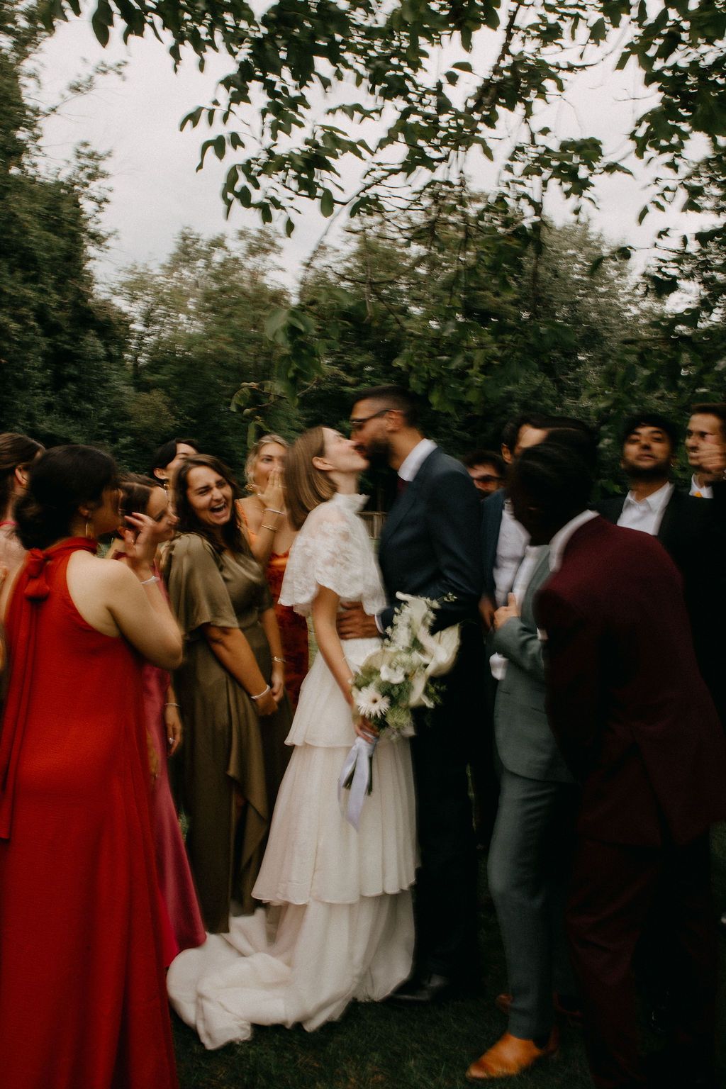 Bride and groom share a kiss surrounded by cheering wedding guests in colorful attire outdoors.
