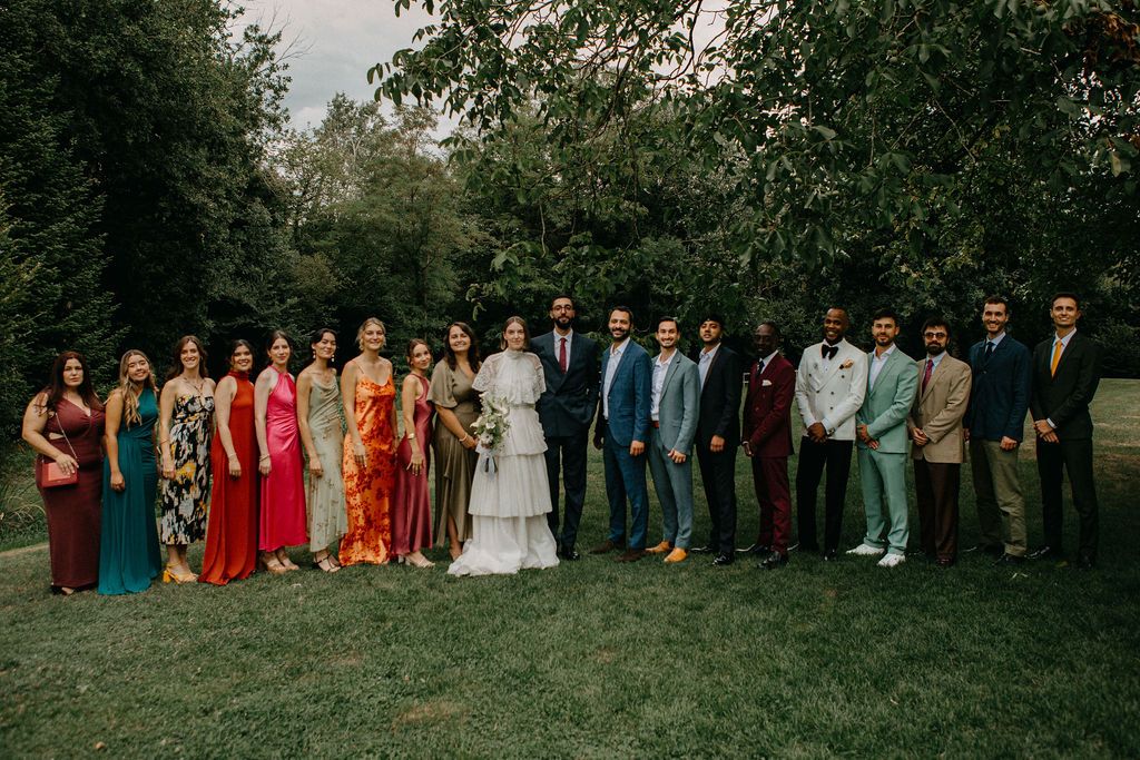 Large wedding party group portrait on green lawn with colorful bridesmaid dresses and suits among trees.