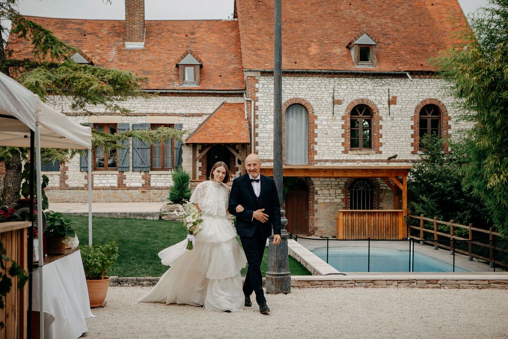 Bride in elegant gown walking with father through charming French countryside wedding venue grounds.