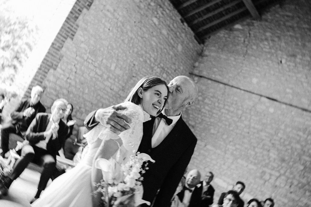 Black and white photo of smiling bride walking down the aisle with her father at stone venue.