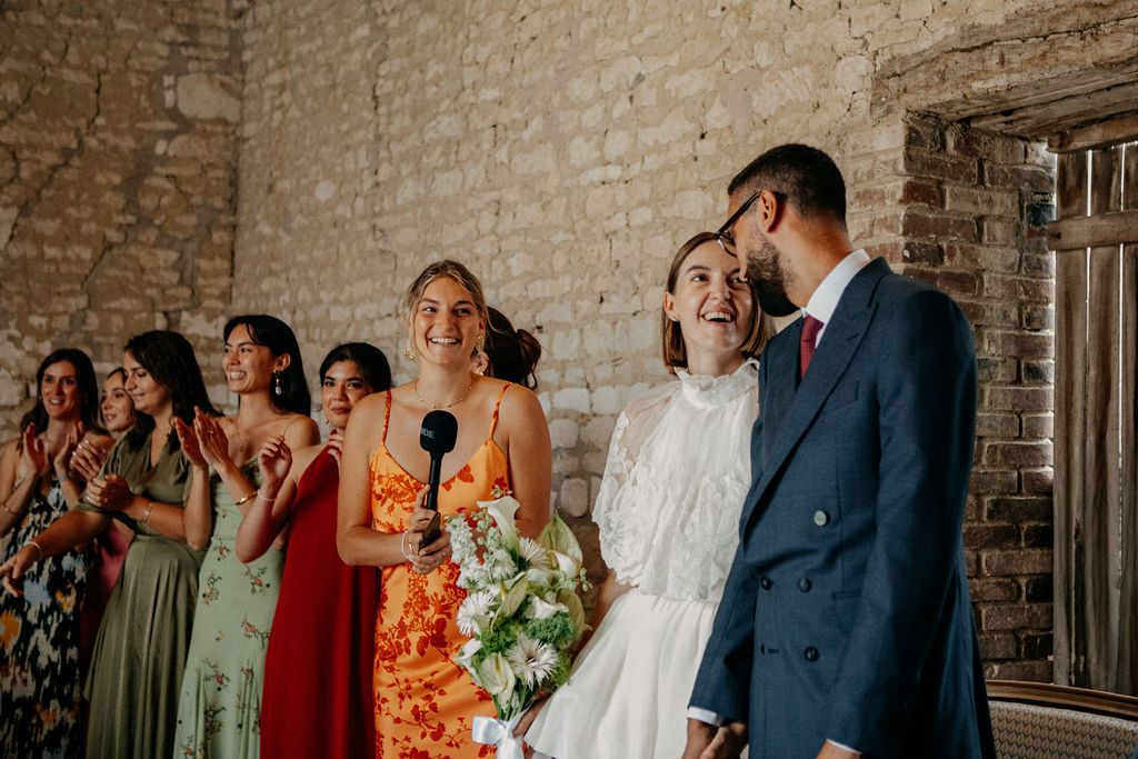 Bride laughing with groom and bridesmaids in colorful dresses during wedding speech at rustic venue.