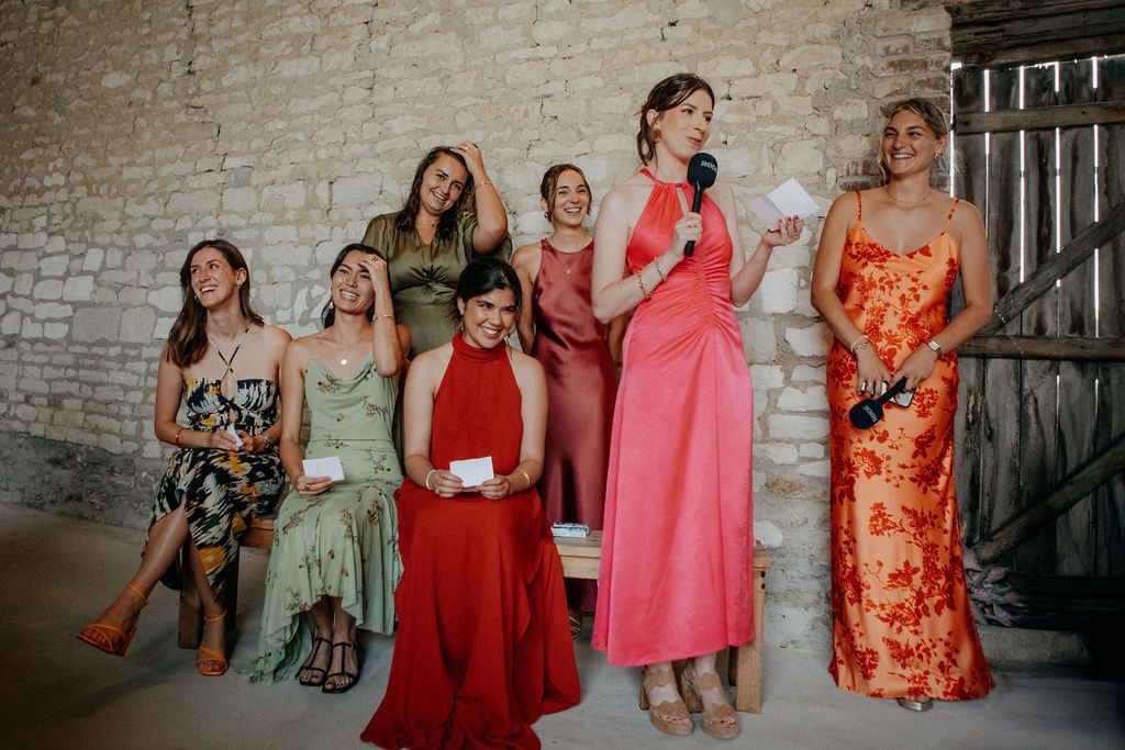 Group of bridesmaids in colorful dresses laughing as one gives a speech with microphone at wedding.