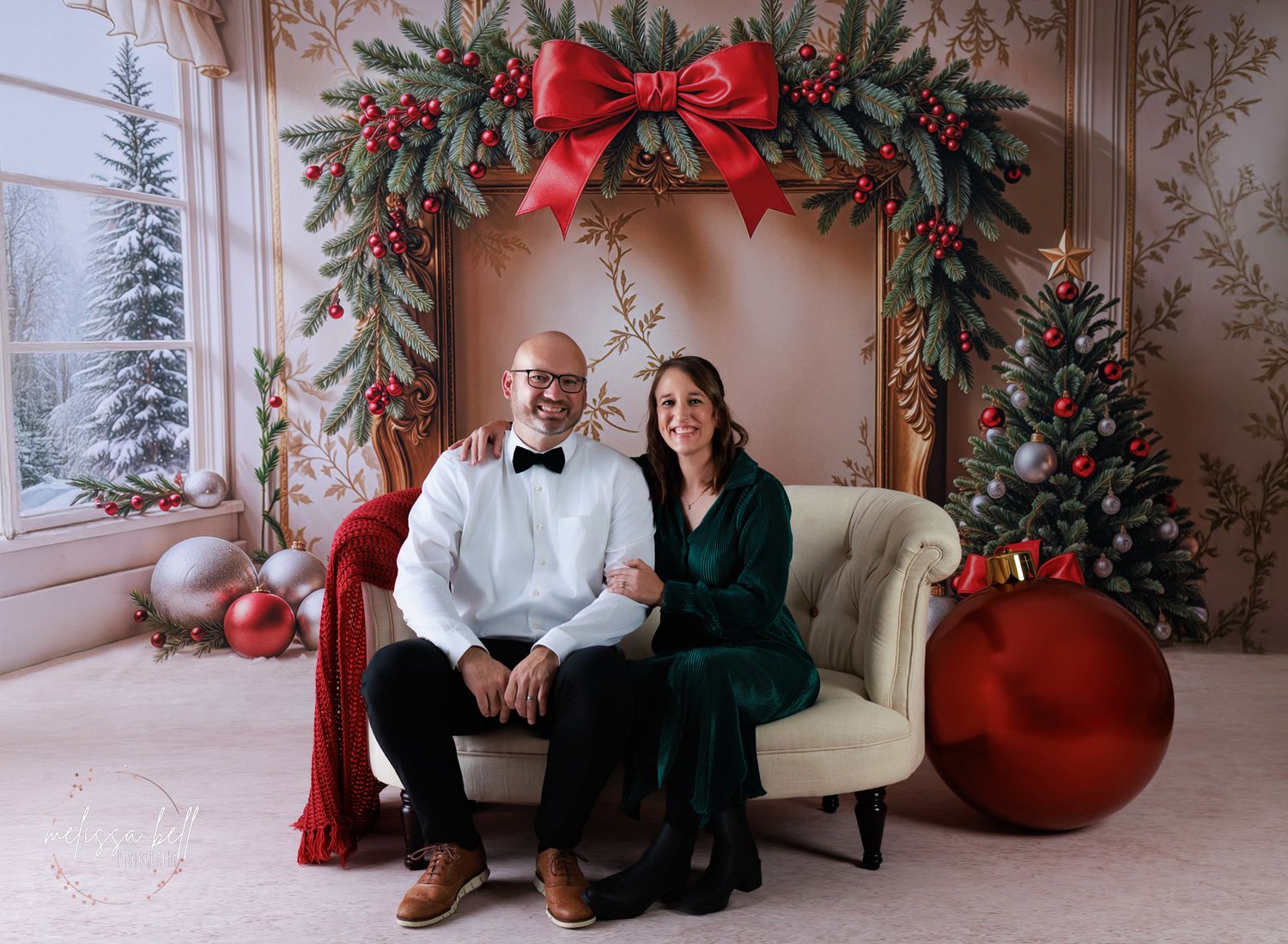 A festive holiday photoshoot against a snowy backdrop with winter decor, red ornaments, and a cream-colored chair.