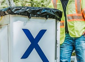 Workers in reflective safety vests stand near white fence panels marked with large blue X symbols at a construction site.