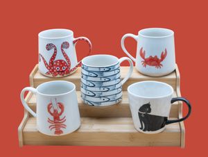 Collection of coastal-themed ceramic mugs and bowls featuring nautical designs of crabs, lobsters and fish on a wooden tray.