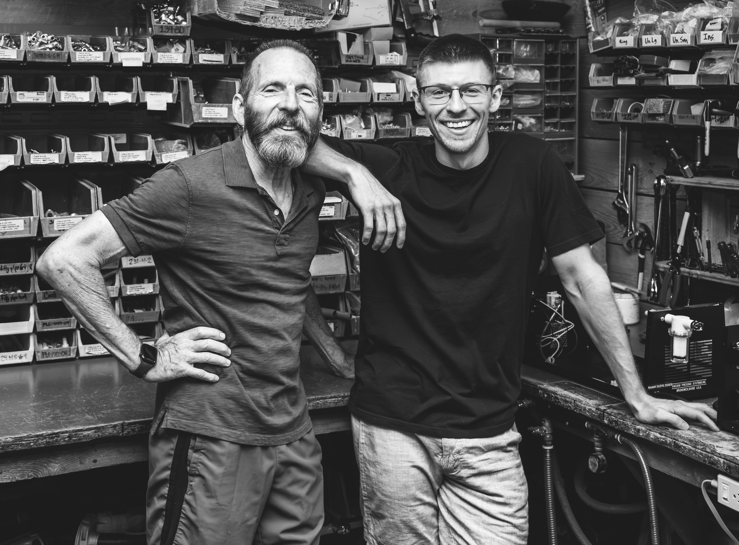 Two people standing together and smiling in what appears to be a workshop or garage with storage shelves in background.