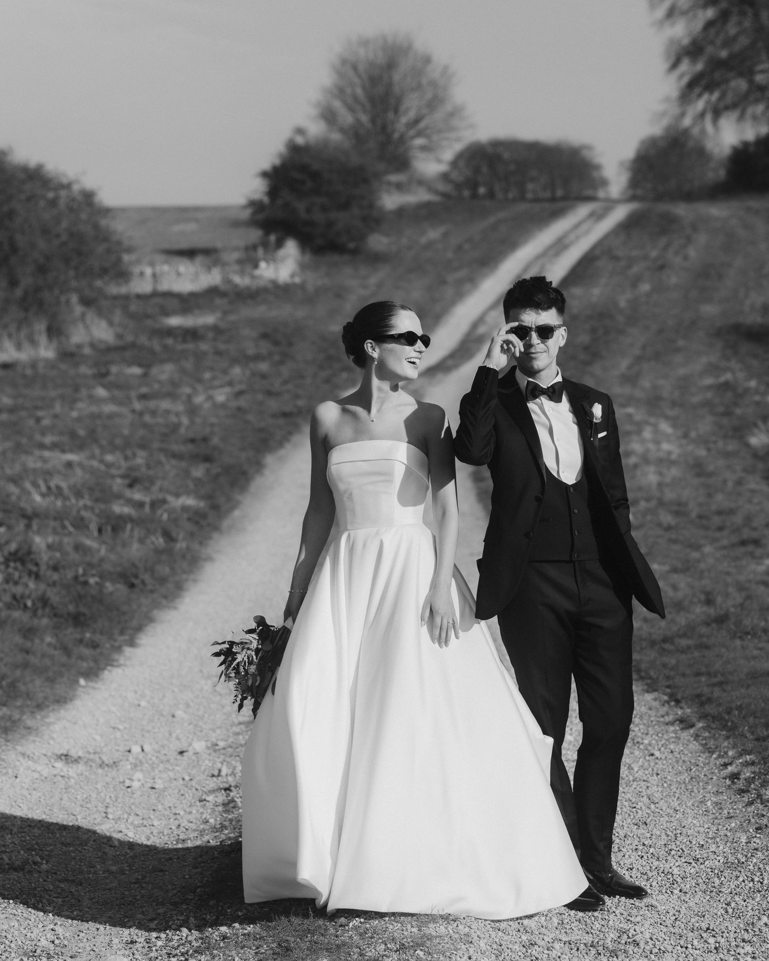 A stylish black and white wedding photo of a couple wearing sunglasses walking on a country dirt road.