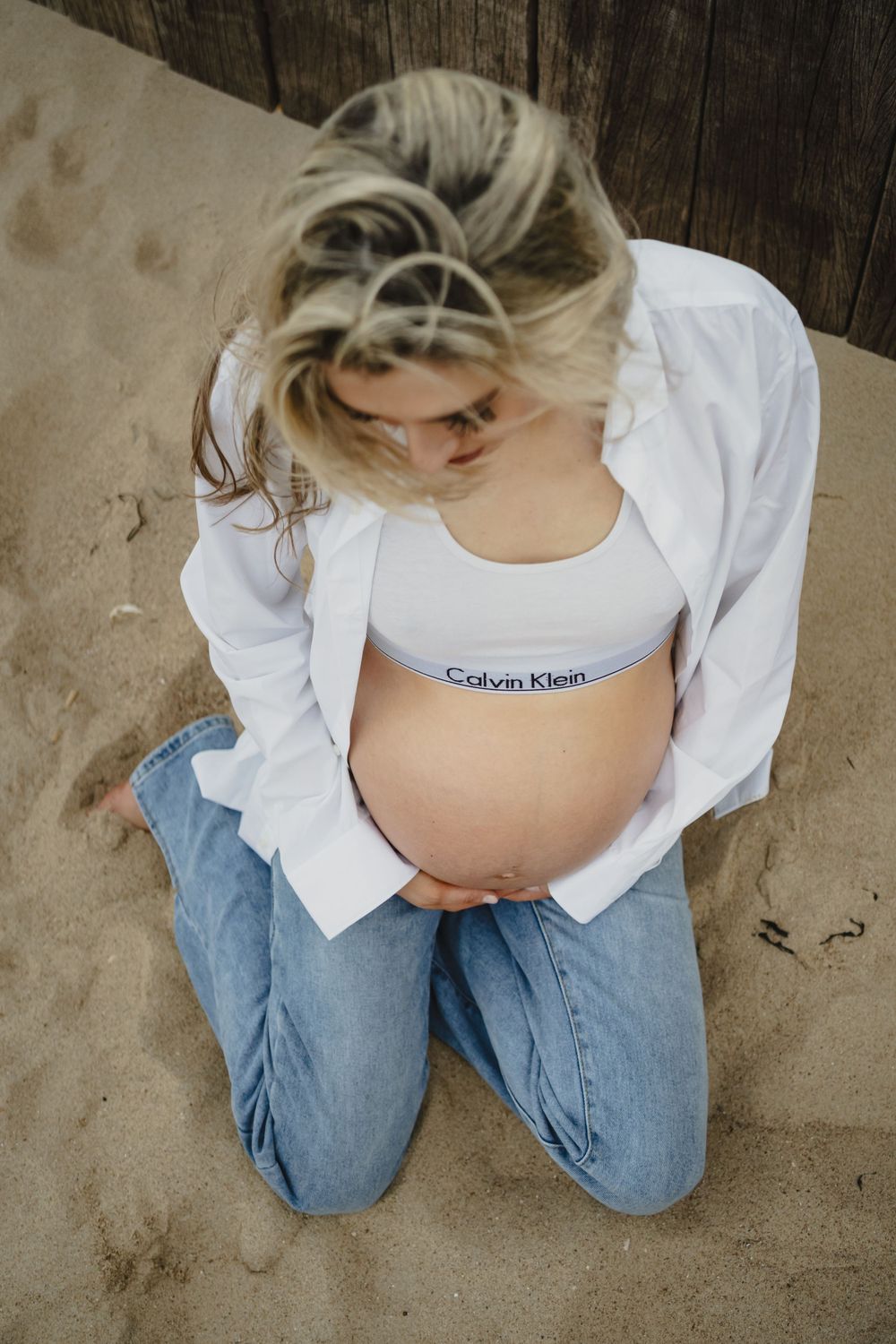 a pregnant mother sat on the beach wearing a white shirt and baggy jeans