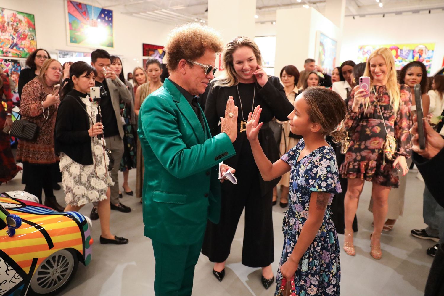 A person in a bright green suit greets a young person at a crowded art gallery opening event.
