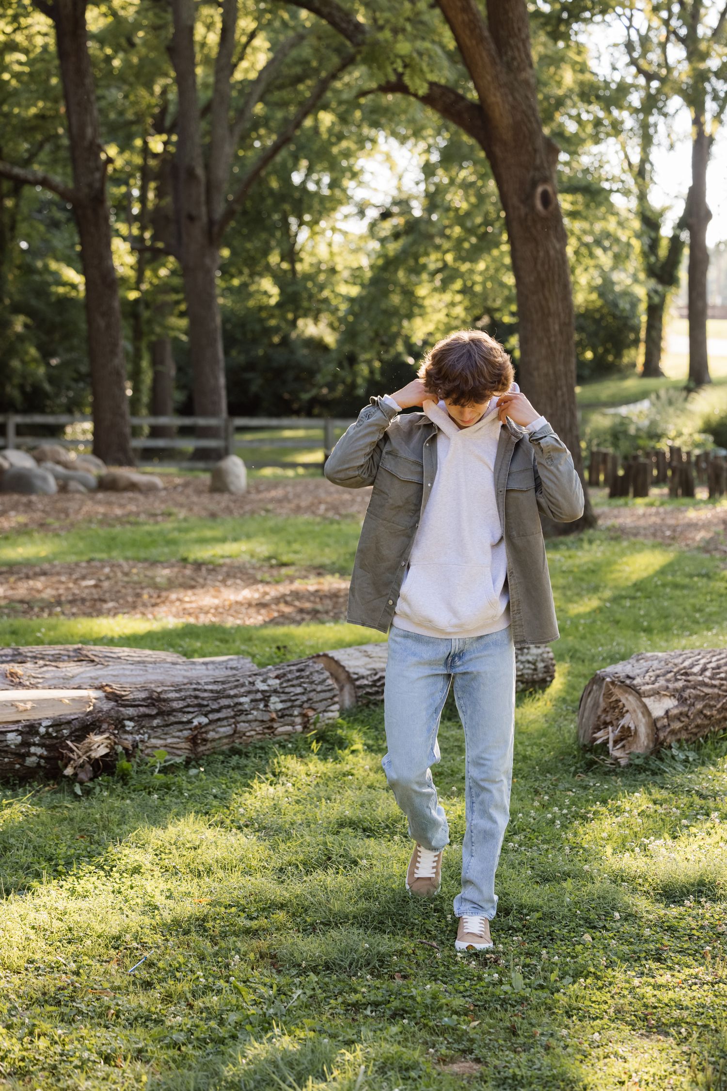 A person in a gray jacket and jeans walking through a sunny park setting.