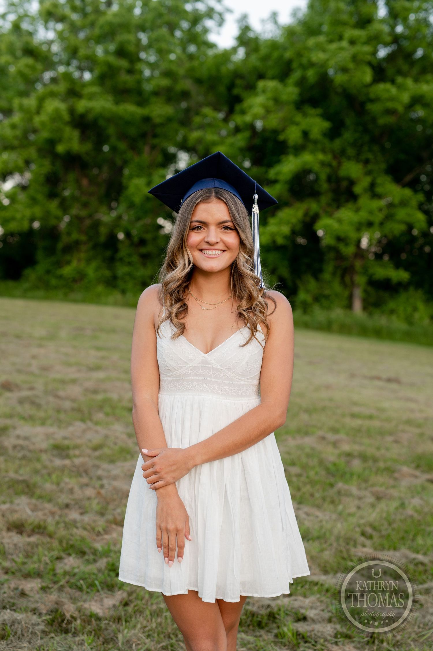 A graduate poses in white dress and navy cap in outdoor setting with green trees.