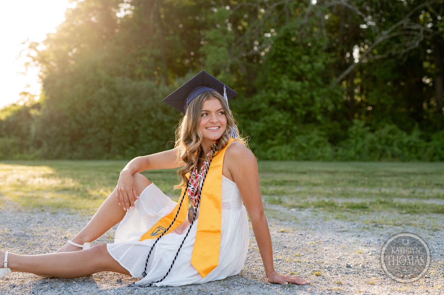 Graduate in white dress and yellow stole sits casually outdoors during sunset photo session.