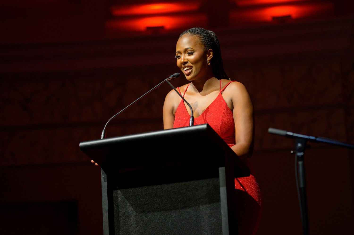 Speaker in red dress presenting at podium during evening event with dramatic lighting.