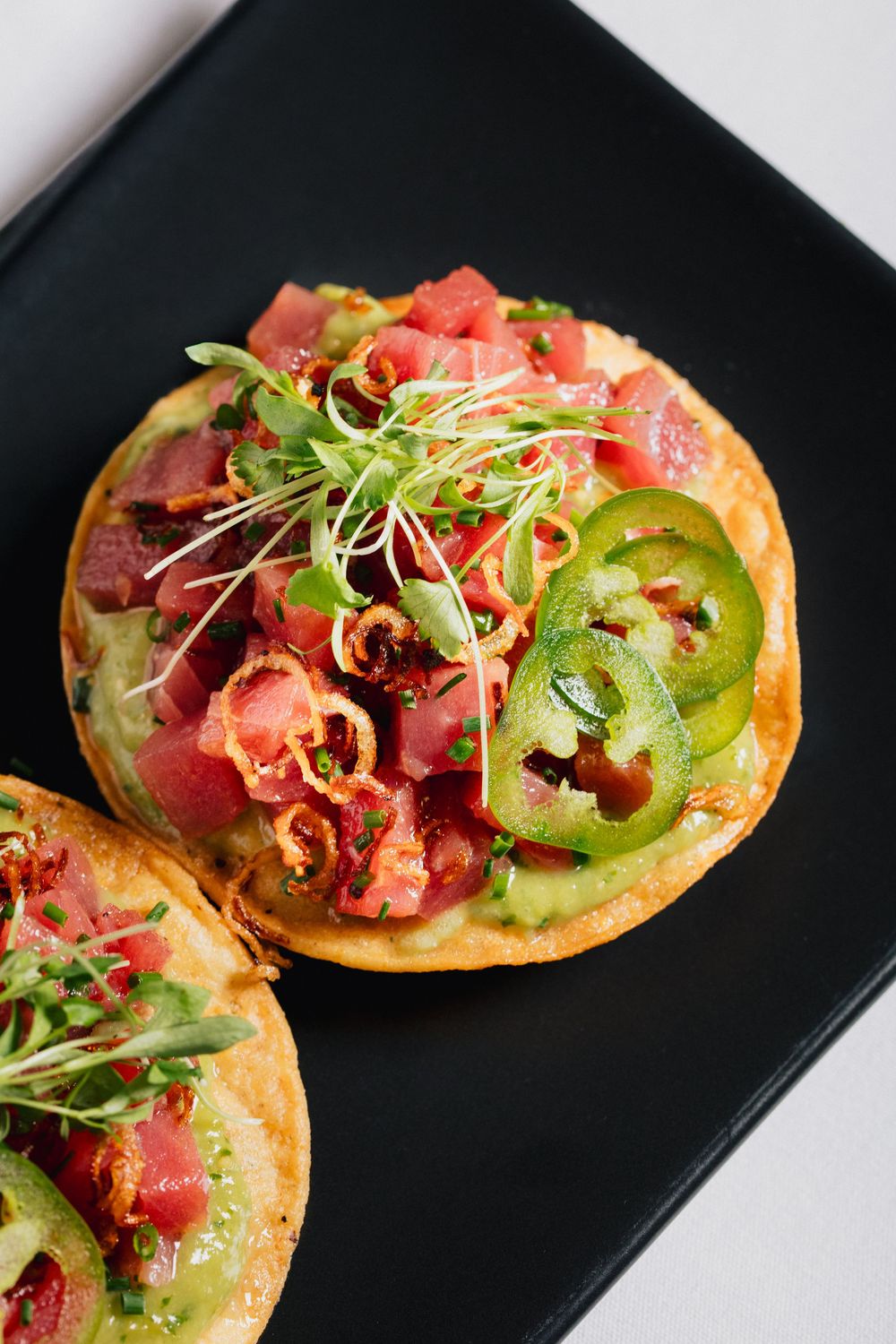 Gourmet tostadas topped with tuna, jalapenos, and microgreens served on a black plate.