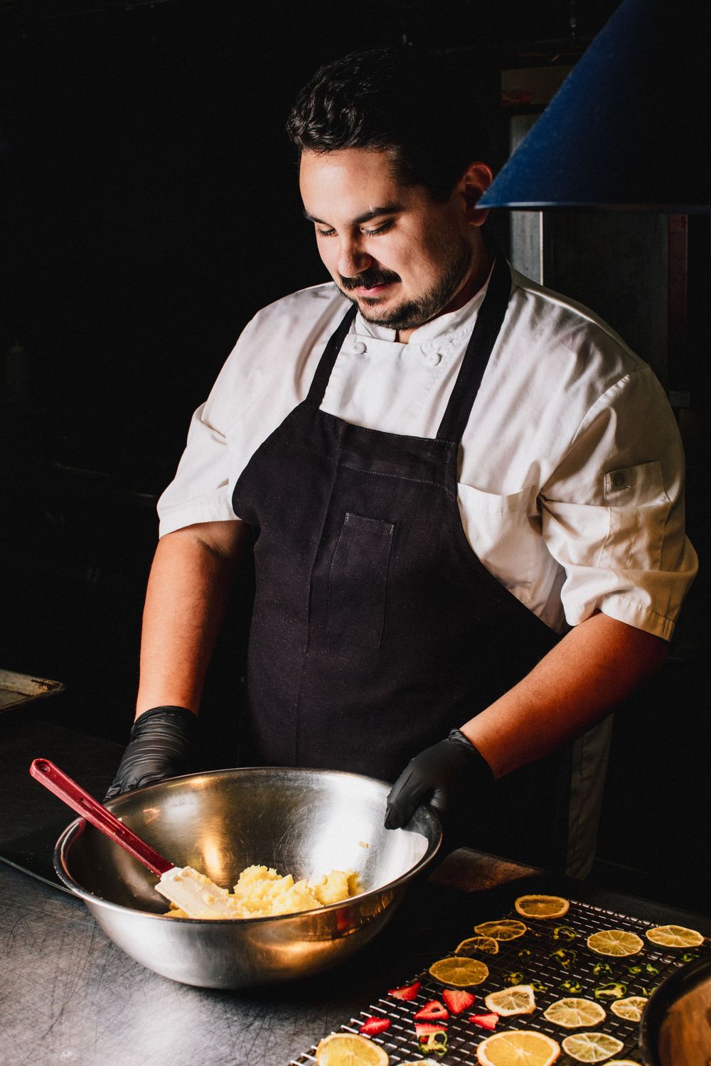 Chef in white uniform and black apron preparing ingredients in a professional kitchen with dramatic lighting.