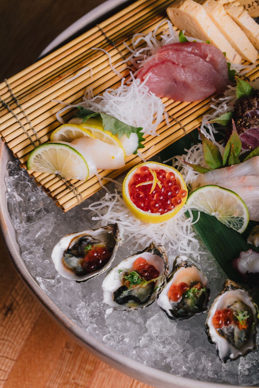 Fresh sashimi and oysters arranged on crushed ice with citrus and roe garnishes on a bamboo mat.