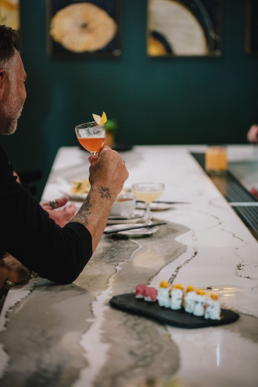 Orange cocktail in coupe glass with sushi rolls served on dark slate at a bar counter.