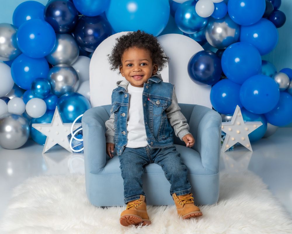 A blue and silver balloon arch with star decorations frames a light blue armchair against a snowy white backdrop.