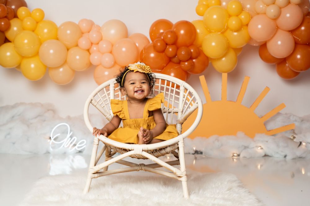 Rattan chair setup with yellow balloon garland arch and orange sun decoration for first birthday photoshoot.