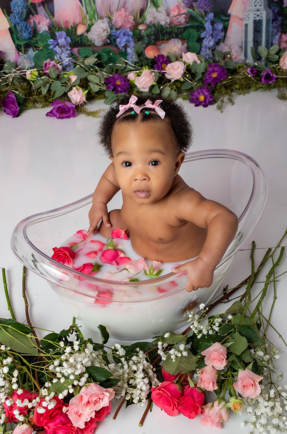 A milk bath photography setup with pink roses and purple flowers surrounding a clear tub with white petals floating in milky water.