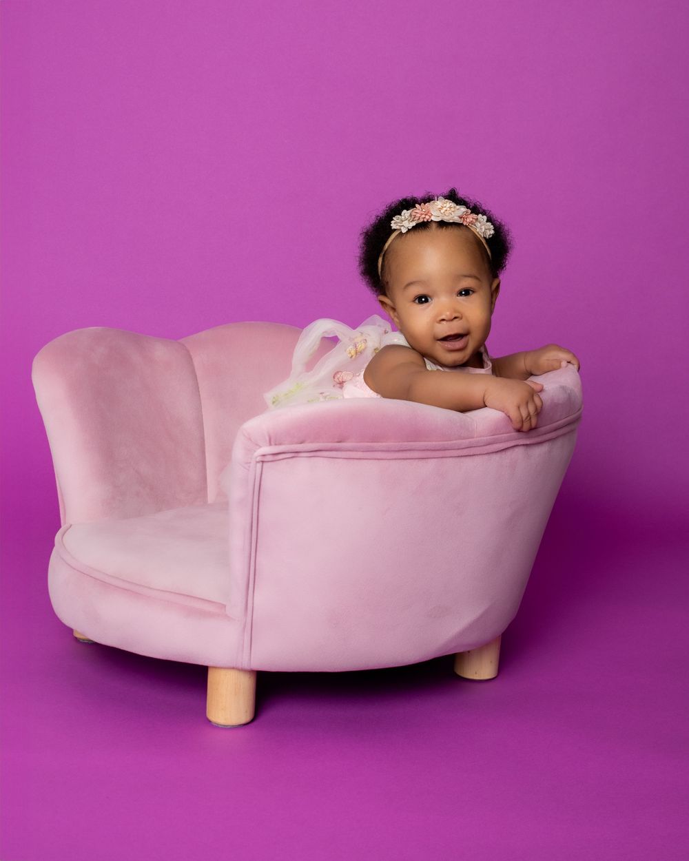 Baby in pink flower crown smiles while posing in light pink chaise lounge against purple backdrop.