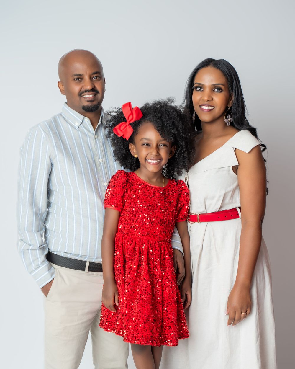 A family of three poses together against a light background, wearing coordinated white and red formal attire.