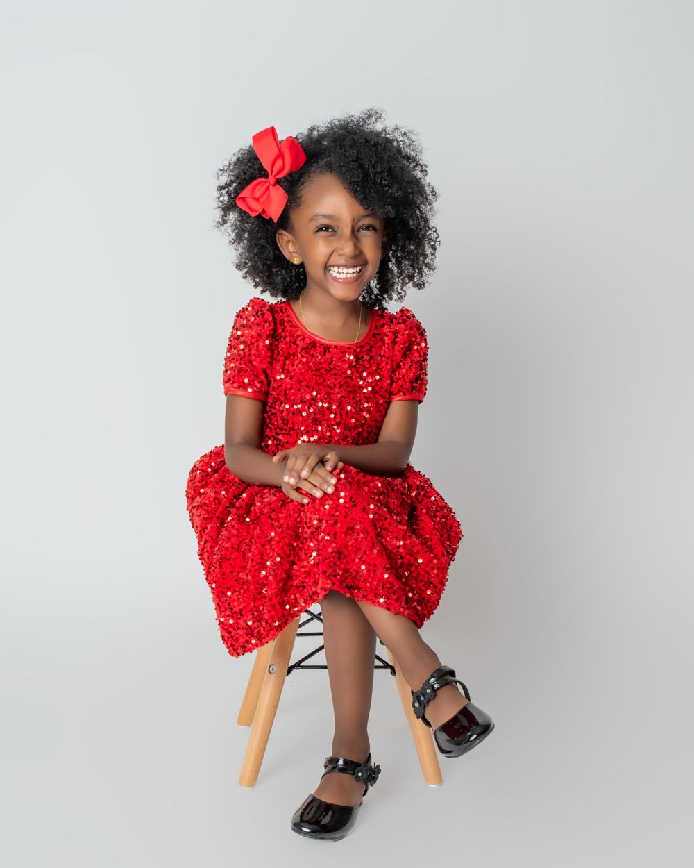 A happy person in a sparkly red holiday dress and red bow sits on a wooden stool against a light gray background.