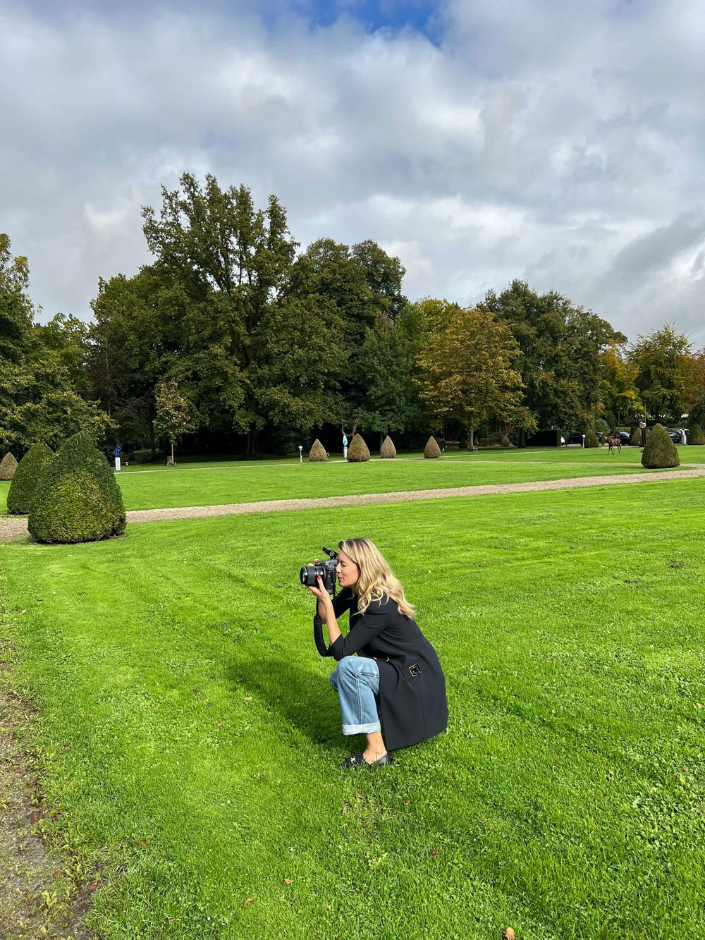 Photographer crouching on manicured green lawn to take picture in garden with ornamental bushes and trees.