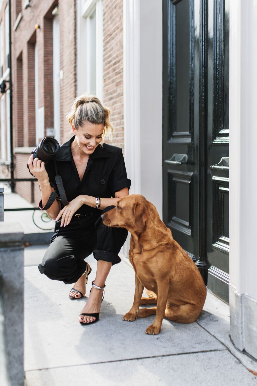 A brown Vizsla dog sits on the sidewalk next to someone in an elegant black outfit and sandals.